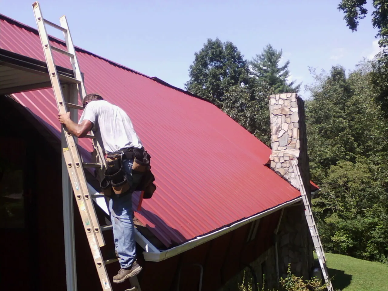 Roofer on a ladder beside a red metal roof and stone chimney — Soffit & Fascia Repair by Roofing Brothers in Huntington Mills